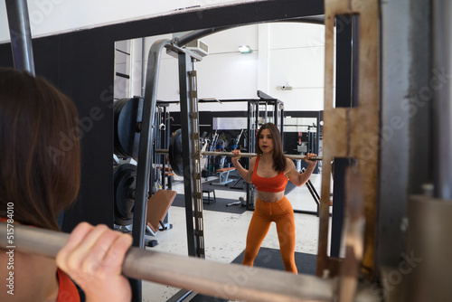 Young and beautiful woman with a sculpted body doing squat exercises with a dumbbell bar on her neck. The woman is wearing orange. Concept Gymnastics, health and wellness.