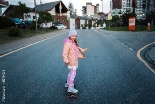 A girl in a pink jacket and hat rides a skateboard on the road
