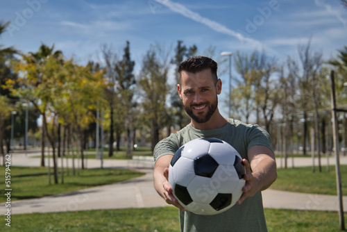 Young and handsome man, with a beard and green shirt, with blue eyes, perfect smile, offering a soccer ball, smiling. Concept sport, ball, football, world, competition. © Manuel