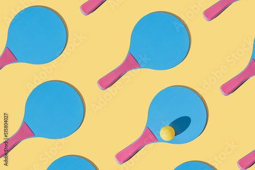 Many blue and pink colourful beach tennis paddle racket with a yellow ping pong ball against yellow background. Flat lay pattern with repetition .Outdoors summer activity, fun relax sport idea.