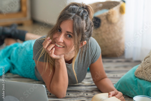 Portrait of happy and cheerful young woman at home laying on the floor and reading a book. Laptop technology computer near her. Pretty female people enjoy indoor leisure activity and relax on sunday photo