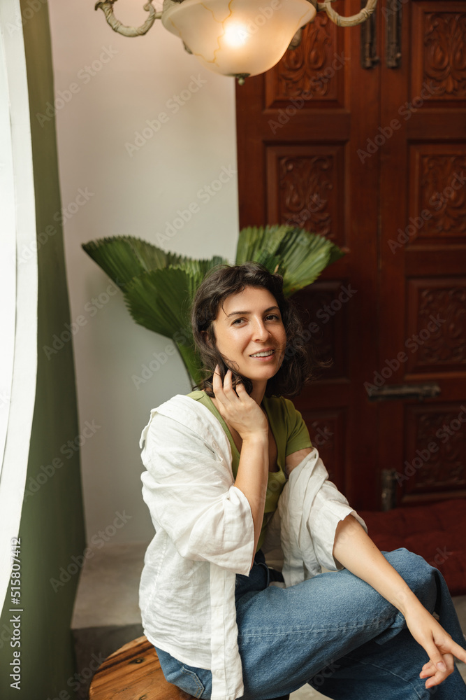 Fototapeta premium Beautiful young caucasian woman is looking at camera, sitting cross-legged indoors. Brunette with wavy hair wears shirt with jeans. Rest time concept.