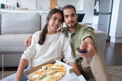Happy couple eating pizza and watching TV. Woman and man sitting at carpet, holding cheese pizza, using remote control. Food, leisure concept