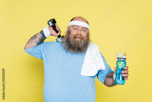 bearded plus size man with towel and sports bottle exercising with dumbbell isolated on yellow Fototapet