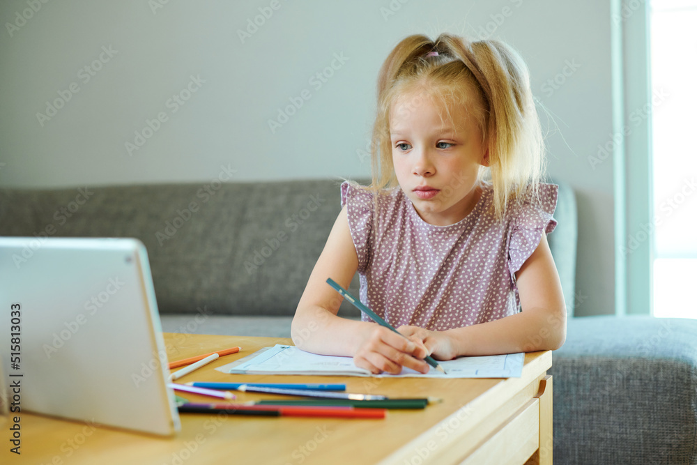 Little child girl learning writing doing homework with digital tablet. Portrait of smart home schooling kid studying alone making note sitting at table. Little child girl learning writing doing homework with digital tablet. Portrait of smart home schooling kid studying alone making note sitting at table.