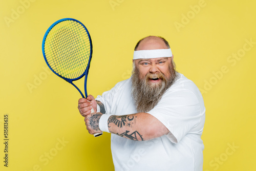 thrilled overweight and tattooed man playing tennis isolated on yellow.