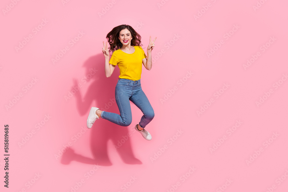 Full length photo of sweet young brunette lady jump show v-sign wear t-shirt jeans shoes isolated on pink background
