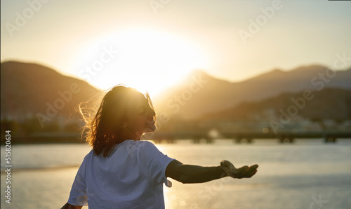 Happy carefree woman turning around to camera with hand, enjoying sunset time with orange sky. Sundown landscape with mountains and sea view