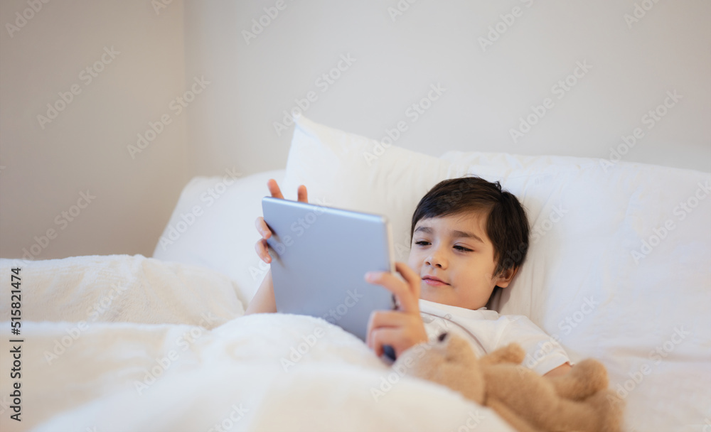 Happy Kid lying in bed holding tablet watching cartoon and chatting with friends on digital pad, Cute young boy playing games online on internet, Child relaxing in the morning before go to school Happy Kid lying in bed holding tablet watching cartoon and chatting with friends on digital pad, Cute young boy playing games online on internet, Child relaxing in the morning before go to school