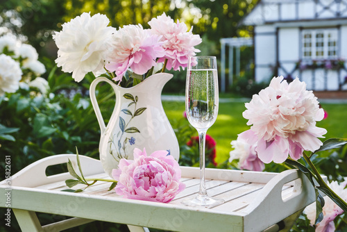 Fototapeta Naklejka Na Ścianę i Meble -  Still life with a glass of champagne or water and bouquet of peonies in jug on a wooden table in a garden. Ready for celebration party in a summer day. Shallow depth of field