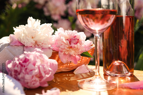 Fototapeta Naklejka Na Ścianę i Meble -  Bottle and glass of rose wine near beautiful peonies on wooden table in garden, closeup
