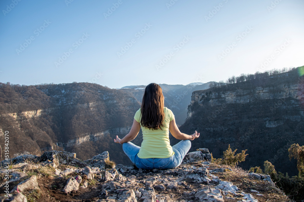 A girl sits on the edge of a cliff in a lotus position, meditates in blue jeans and a yellow T-shirt, view from the back