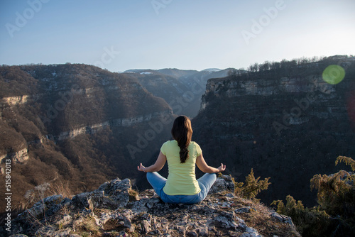 A girl sits on the edge of a cliff in a lotus position  meditates in blue jeans and a yellow T-shirt  view from the back