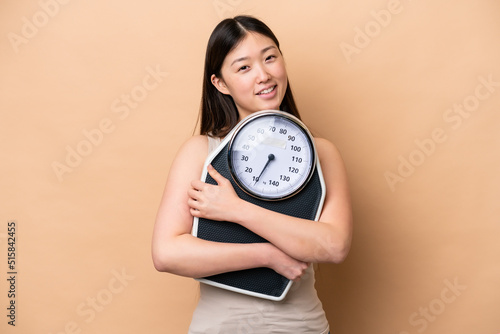 Young Chinese woman isolated on beige background with weighing machine