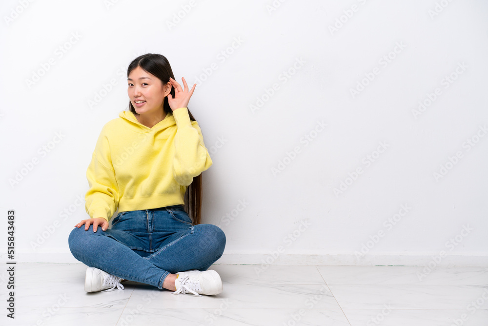 Young Chinese woman sitting on the floor isolated on white wall listening to something by putting hand on the ear Young Chinese woman sitting on the floor isolated on white wall listening to something by putting hand on the ear
