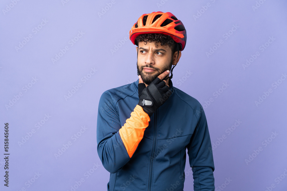 Young Moroccan cyclist man isolated on purple background having doubts - obrazy, fototapety, plakaty 