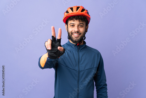 Young Moroccan cyclist man isolated on purple background smiling and showing victory sign