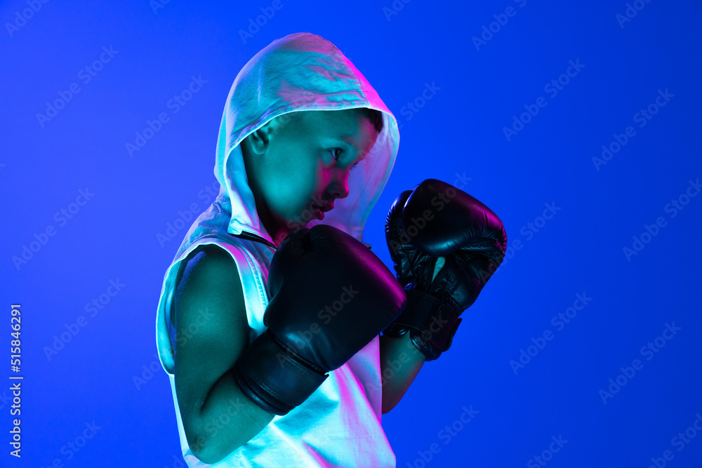 Stylish boy, beginner boxer training isolated over blue background in neon light. Concept of sport, fashion, studying, achievements, skills.