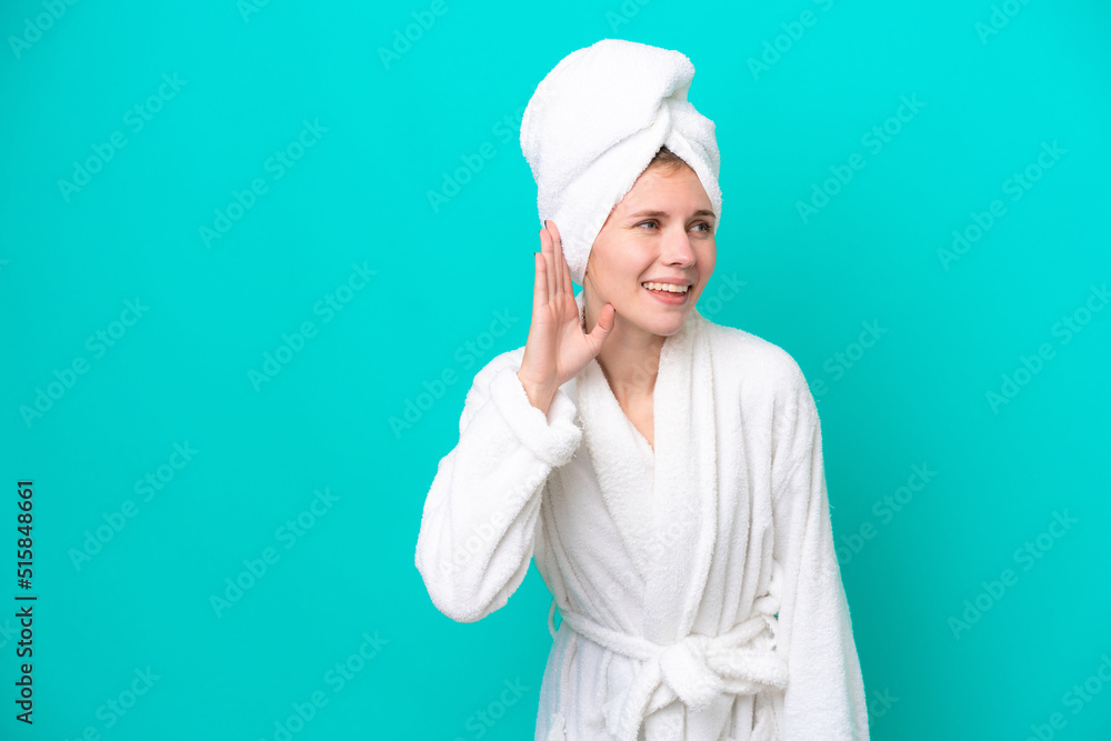 Young woman in a bathrobe isolated on blue background listening to something by putting hand on the ear Young woman in a bathrobe isolated on blue background listening to something by putting hand on the ear