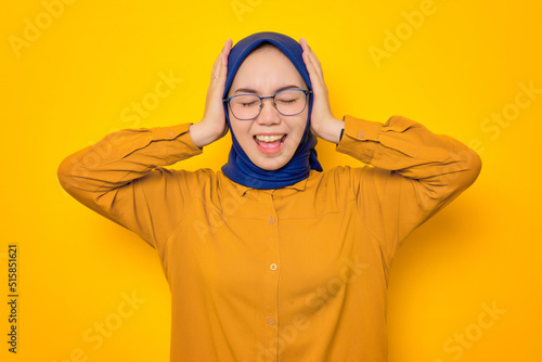 Angry young Asian Muslim woman dressed in orange covering ears with hands and screaming loudly isolated on yellow background © Sewupari Studio