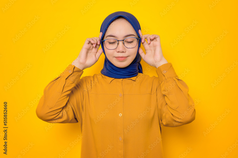 Attractive young Asian Muslim woman dressed in orange shirt holding fingers on temples, trying to concentrate isolated on yellow background