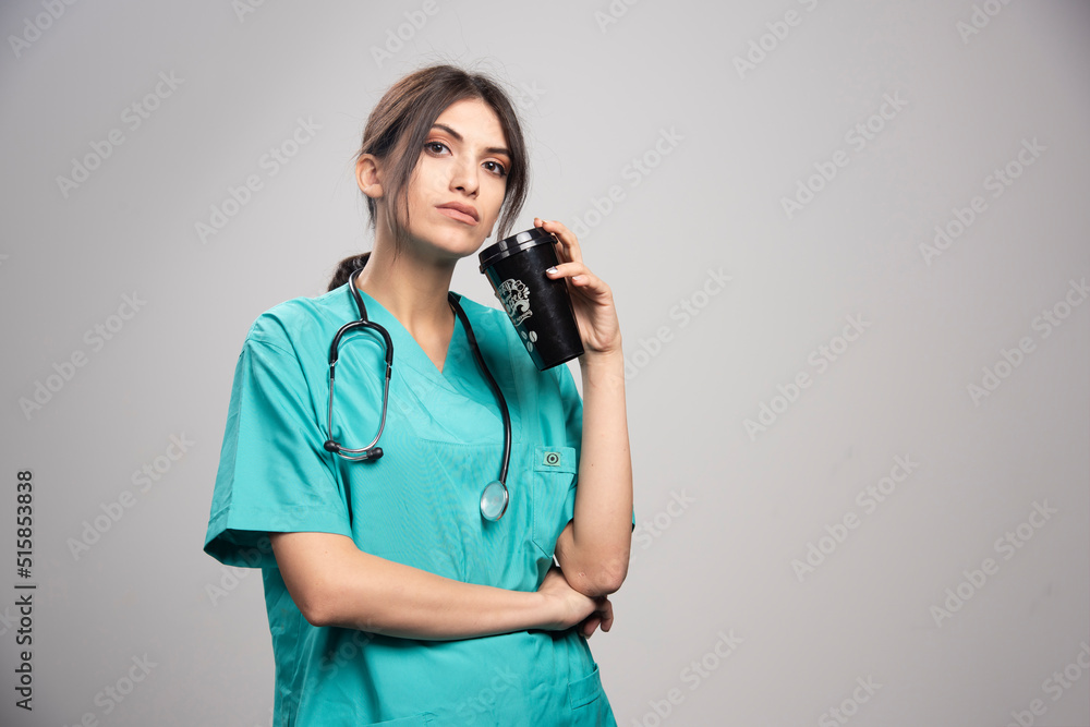 Female doctor posing with cup of coffee on gray background Female doctor posing with cup of coffee on gray background