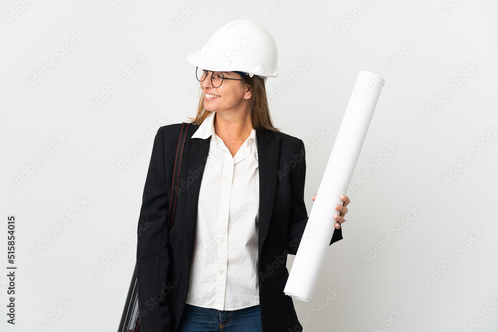 Middle age architect woman with helmet and holding blueprints over isolated background looking to the side and smiling Middle age architect woman with helmet and holding blueprints over isolated background looking to the side and smiling