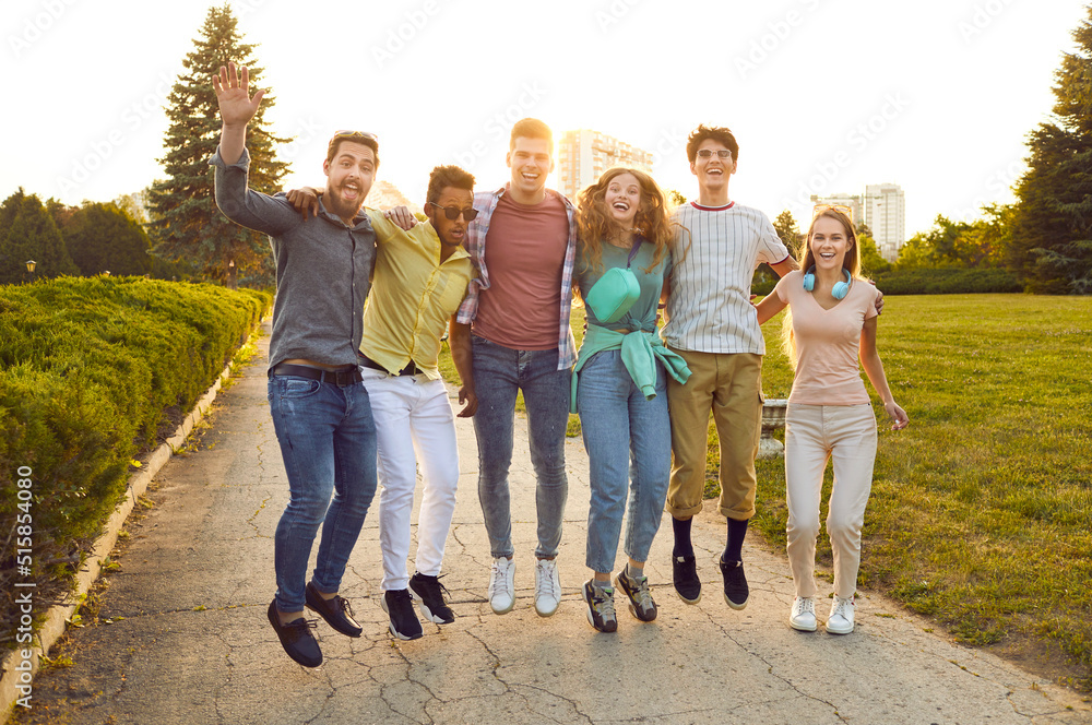 Fun group of best friends hugging and jumping up in park enjoying summer weekend. Young multiracial men and women in youth casual clothes standing in row bouncing on walkway in city public park.
