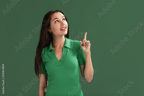 Portrait of beautiful young girl posing, rising finger, having idea isolated over green studio background
