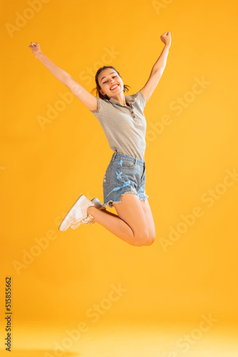 Full-length portrait of beautiful young girl in casual outfit jumping, posing isolated over yellow studio background