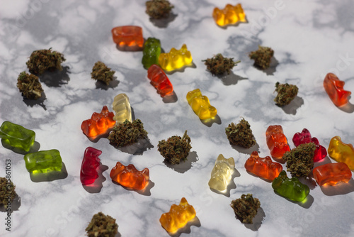 Several dry buds of medical marijuana lie at random among gummy bears of different colors with the addition of cbd oil on a marble table photo