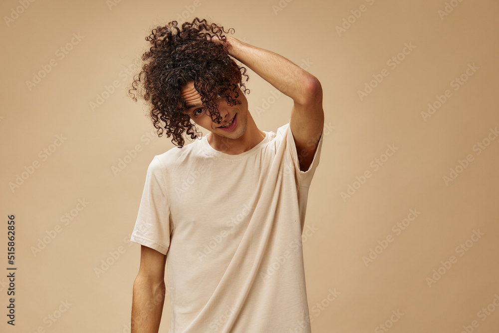 Funny awesome tanned curly man in basic t-shirt touches hair curls posing isolated on over beige pastel background. Fashion New Collection offer. People and Emotions concept. Free place for ad Funny awesome tanned curly man in basic t-shirt touches hair curls posing isolated on over beige pastel background. Fashion New Collection offer. People and Emotions concept. Free place for ad