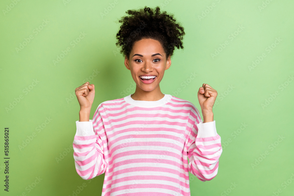 Photo of lucky sweet woman wear striped sweater smiling rising fists isolated green color background Photo of lucky sweet woman wear striped sweater smiling rising fists isolated green color background