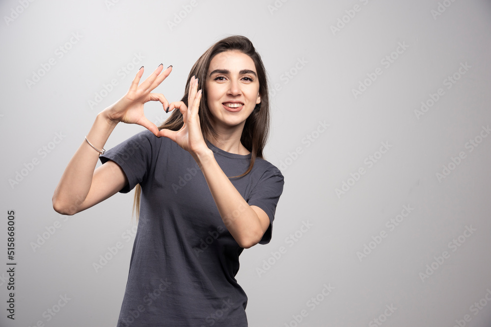 Young woman wearing gray t-shirt over gray background doing heart symbol shape with hands Young woman wearing gray t-shirt over gray background doing heart symbol shape with hands