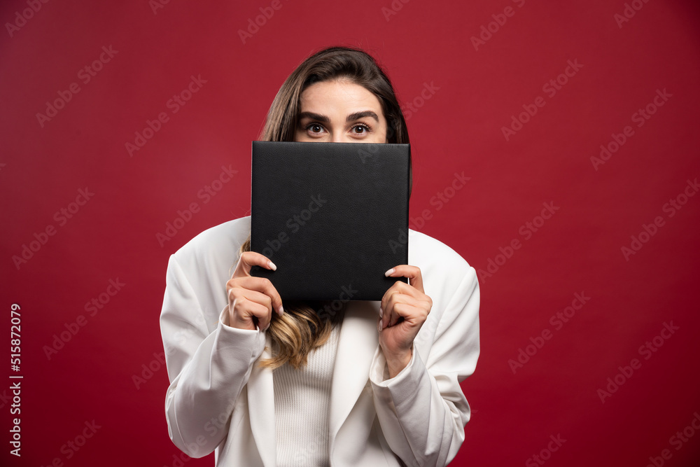 Business woman covering her face with a notebook on a red background Business woman covering her face with a notebook on a red background