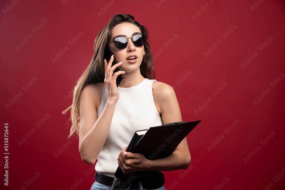 Young pretty woman with glasses speaking on phone and holding notebooks on a red background Young pretty woman with glasses speaking on phone and holding notebooks on a red background