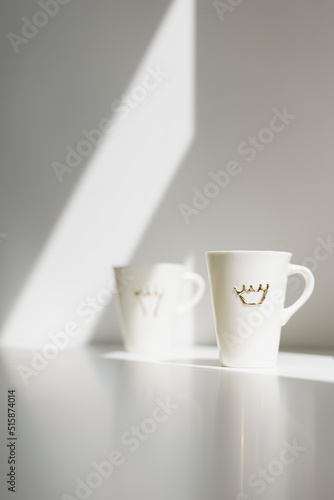 Two white tall porcelain coffee cups with crowns on a white table with sun hitting the wall behind creating a nice morning atmosphere