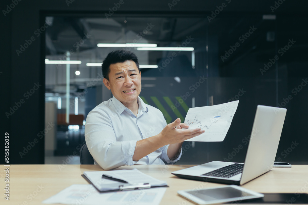 An Asian broker is disappointed with the company's financial indicators, looks at the camera not happy, shows a document with a negative income schedule, the man works in the office. An Asian broker is disappointed with the company's financial indicators, looks at the camera not happy, shows a document with a negative income schedule, the man works in the office.