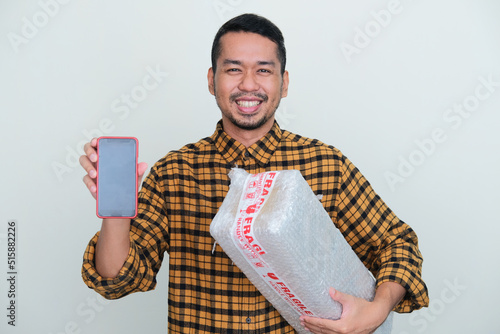 Adult Asian man smiling while showing blank mobile phone screen and holding wrapped package photo