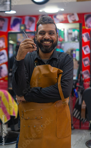 Attractive barber positive arab man smiling for camera and showing hairdressing scissors while working in vintage salon against barbershop background photo