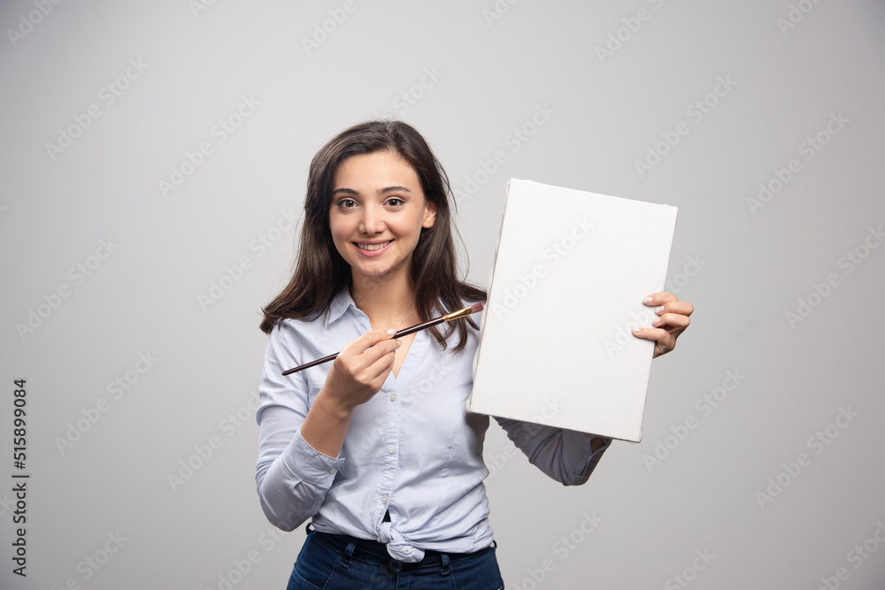 Brunette painter posing with canvas and brush on gray background Brunette painter posing with canvas and brush on gray background