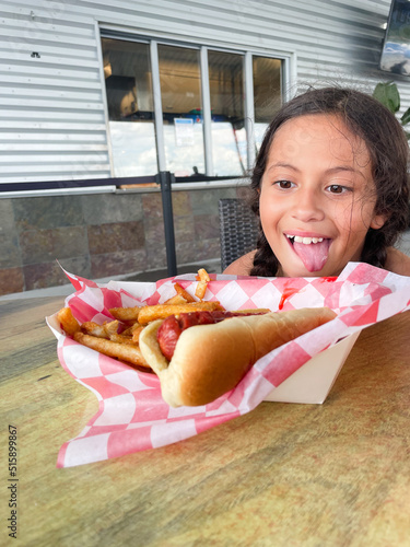 young girl sitting and staring hungrily at fresh hot dog and fries with tongue out delicious meal outdoor summer fun