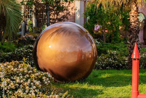 A large bronze-colored glossy ball on a green lawn among flowers and southern plants in the park. Garden decor. Landscaping © jockermax3d