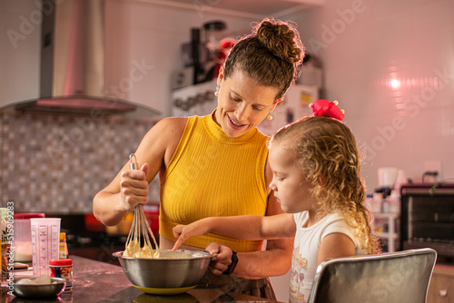 Imagem de mãe e filha brincando enquanto cozinham um bolo. Mulher loira brincando com sua filha na cozinha. Receita de doce feita em família.