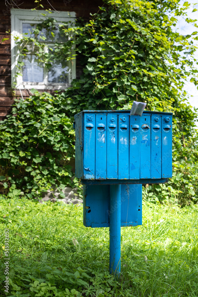 An old blue iron mailbox for mail and correspondence in a village near a house overgrown with greenery.