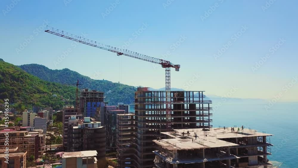 Drone view of construction site, multi-storey buildings under construction against backdrop of sea and mountains on sunny summer day. Construction equipment, crane and workers walk around building. Drone view of construction site, multi-storey buildings under construction against backdrop of sea and mountains on sunny summer day. Construction equipment, crane and workers walk around building.