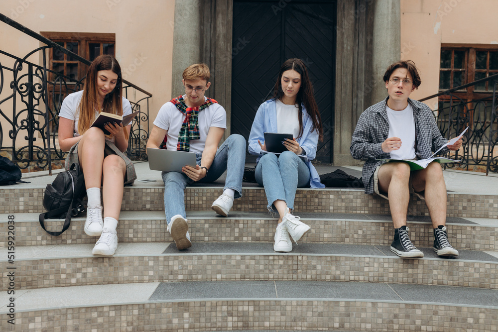 Fototapeta premium Study together is fun, teamwork, team building concept. Four happy students are sitting near college building and holding books, devices, wearing casual smart, smiling, on a nice summer
