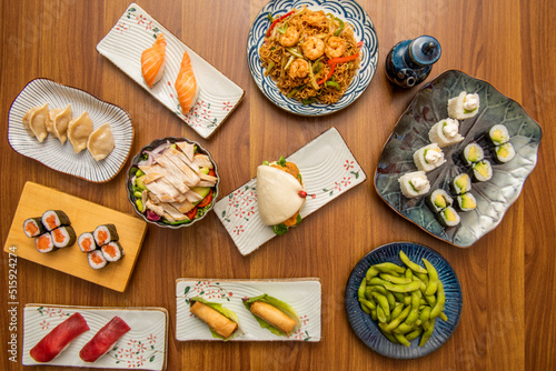 set of asian food dishes with sushi, avocado maki, shrimp noodles, soy sauce, spring rolls, bluefin tuna, edamame beans, gyozas, norwegian salmon and bao bread on brown wooden table