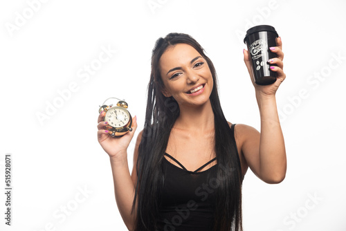 Happy young woman with cup holding an alarm clock on white background
