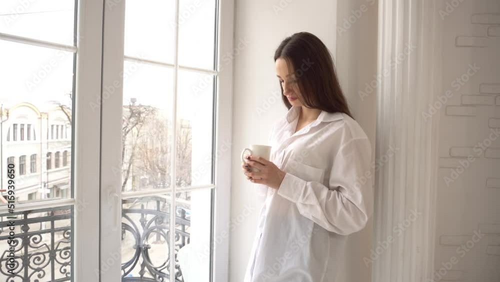 4k young woman standing by window drinking from coffee cup 4k young woman standing by window drinking from coffee cup